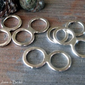 May include: Silver-toned metal jewellery findings with two connected rings. The findings are arranged in a pile on a wooden surface.