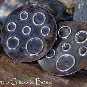 May include: Two copper pendants with a hammered finish and a circle design. The circles are outlined in silver.