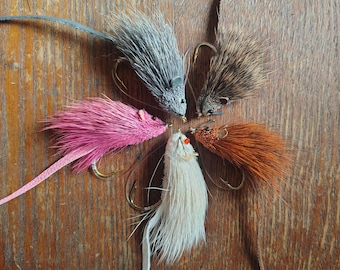 May include: Five mouse-shaped fishing lures arranged in a circle. The lures feature fur bodies in gray, brown, pink, white, and rust orange, each with a metal hook, small bead eyes, and a long, thin leather tail extending from the back of the body.