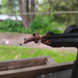 May include: A brown fuzzy fishing fly lure featuring dark beads and thin fibres, secured in a black metal vice. The lure has a textured body with protruding hair-like elements and small metallic spheres attached to the front section.