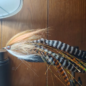 May include: A fishing fly lure featuring a mix of tan, white, and orange synthetic fibres, accented with long, black and white barred feathers and iridescent gold tinsel strands. The lure includes a small, reflective copper-coloured eye detail.