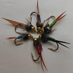 May include: A collection of five fishing flies arranged in a circular pattern. Each fly features a gold-coloured metal bead head, a dark feathered thorax, and a wire-wrapped body in metallic copper, blue, red, and green colours with long tails.