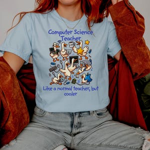 Puede incluir: Camiseta azul claro con el texto "Computer Science Teacher" y "Like a normal teacher, but cooler." La camiseta presenta un diseño de dibujos animados de gansos con portátiles, estrellas y accesorios informáticos.