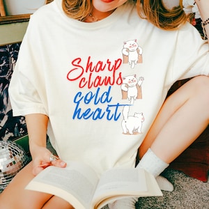 Può includere: T-shirt color crema con la scritta "Sharp claws, gold heart" in corsivo rosso e blu. La maglietta presenta tre gatti dei cartoni animati in varie pose. Un capo casual per tutti i giorni.