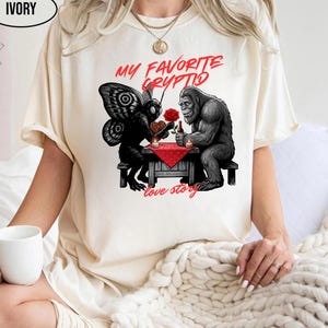 Puede incluir: Camiseta de color marfil con un gráfico de una polilla y un gorila en una mesa, con el texto "MY FAVORITE CRYPTID" y "love story" en rojo. La polilla sostiene un corazón y el gorila sostiene una rosa.