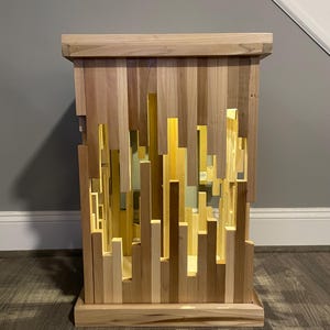 May include: A wooden end table with a unique design featuring a geometric pattern of vertical slats. The slats are arranged in a random pattern, creating a modern and abstract look. The table is made of light wood and has a natural finish.