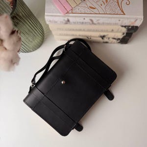 K&ouml;nnte beinhalten: Eine schwarze Leder-Umh&auml;ngetasche mit Klappenverschluss und einer silbernen Schnalle. Die Tasche hat einen verstellbaren Riemen, der bequem auf der Schulter getragen werden kann.