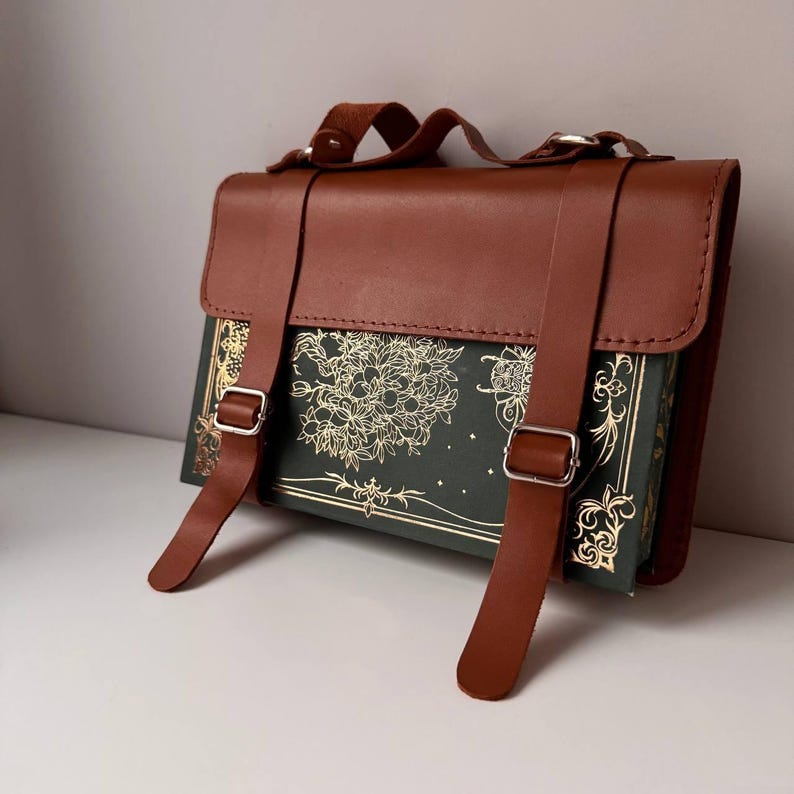 K&ouml;nnte beinhalten: Eine braune Leder-Buch-Tasche mit einem gr&uuml;nen Buchumschlag, der mit einem goldenen Blumenmuster verziert ist. Die Tasche hat zwei verstellbare Tr&auml;ger mit silbernen Schnallen.