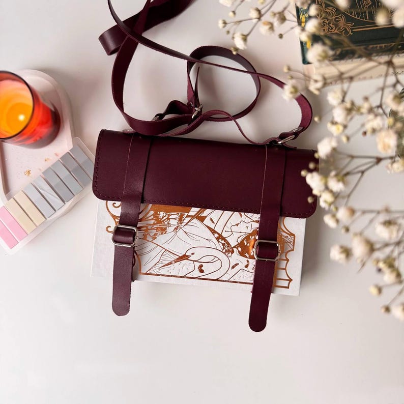 K&ouml;nnte beinhalten: Eine bordeauxfarbene Leder-Umh&auml;ngetasche mit einem wei&szlig;en und goldenen Design. Die Tasche hat zwei verstellbare Tr&auml;ger mit silbernen Schnallen.