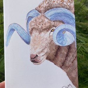 May include: Watercolor painting of a brown sheep with large blue horns. The sheep's eye is visible and the painting is signed in the bottom right corner.