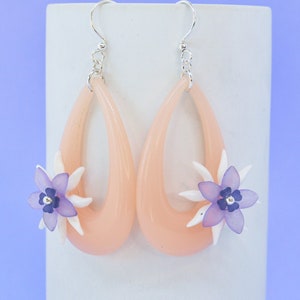 May include: A pair of peach-colored dangle earrings with a delicate flower design. The flowers are made of white and purple polymer clay and have a silver center.