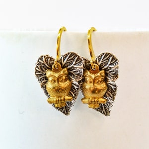 May include: Gold owl earrings with silver leaf details. The owls are perched on a branch and the leaves are arranged in a heart shape.