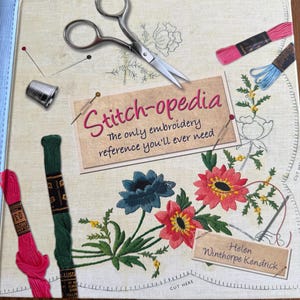 May include: An open book titled "Stitch-opedia" with embroidery supplies. The book features the text "The only embroidery reference you'll ever need." and the author's name. Embroidery floss, scissors, and a thimble are also visible.