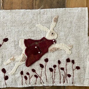 May include: A white rabbit wearing a red dress jumps over a field of burgundy flowers. The rabbit is made of felt and the flowers are embroidered on a beige linen background.