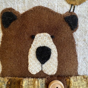 May include: A brown felt bear with a white felt nose and black felt eyes. The bear is wearing a brown felt scarf with a plaid pattern and two wooden buttons. A small brown felt bird with a yellow beak is perched on top of the bear's head.