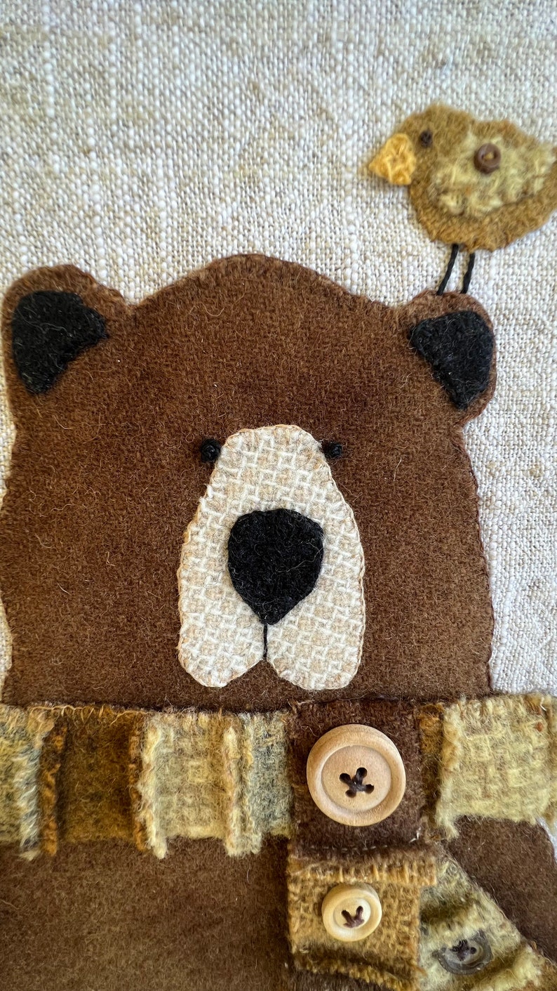 May include: A brown felt bear with a white felt nose and black felt eyes. The bear is wearing a brown felt scarf with a plaid pattern and two wooden buttons. A small brown felt bird with a yellow beak is perched on top of the bear's head.