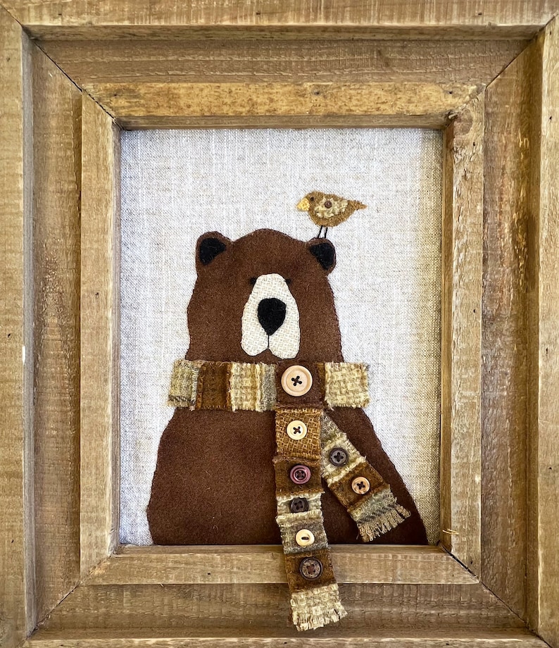 May include: A brown felt bear wearing a brown, tan and white striped scarf with buttons. A small brown bird sits on top of the bear's head. The artwork is framed in a rustic wooden frame.