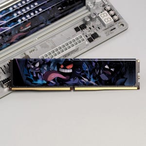 May include: A computer RAM memory module featuring a dark purple and blue graphic print of the character Gengar with a long tongue. The module has gold-colored contact pins along the bottom edge and is positioned over a computer motherboard.