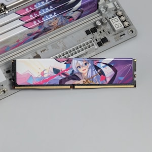 May include: A computer RAM memory module featuring a printed graphic of an anime character with long light blue hair and a wand. The module has a black base with gold connector pins and is positioned over a gray motherboard with additional RAM slots.