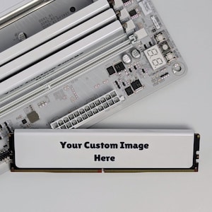 K&ouml;nnte beinhalten: Ein wei&szlig;es Computer-Mainboard mit silbernen RAM-Steckpl&auml;tzen und einer digitalen Debug-Anzeige, neben einem einzelnen RAM-Riegel mit wei&szlig;er Oberfl&auml;che und dem schwarzen Aufdruck "Your Custom Image Here" auf dem K&uuml;hlk&ouml;rper des Moduls.