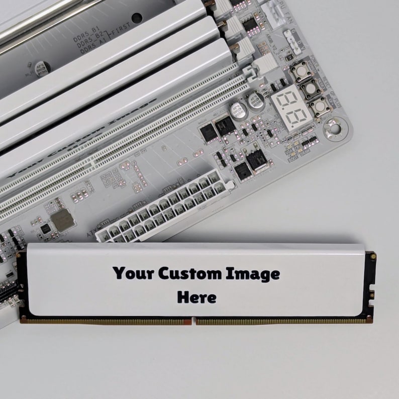 K&ouml;nnte beinhalten: Ein wei&szlig;es Computer-Mainboard mit silbernen RAM-Steckpl&auml;tzen und einer digitalen Debug-Anzeige, neben einem einzelnen RAM-Riegel mit wei&szlig;er Oberfl&auml;che und dem schwarzen Aufdruck "Your Custom Image Here" auf dem K&uuml;hlk&ouml;rper des Moduls.