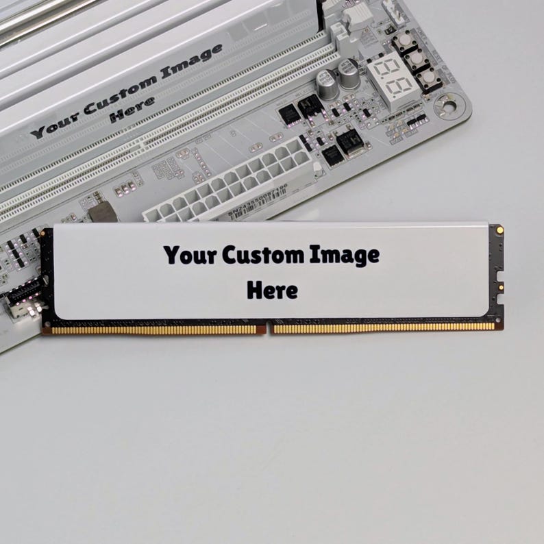 K&ouml;nnte beinhalten: Ein wei&szlig;es RAM-Speichermodul mit einem leeren rechteckigen Bereich, der den schwarzen Text Your Custom Image Here enth&auml;lt, platziert vor einem Computer-Mainboard mit einem passenden wei&szlig;en K&uuml;hlk&ouml;rper und demselben aufgedruckten Text.