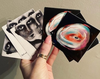 Puede incluir: Un conjunto de pegatinas con arte abstracto sobre fondo negro. Una pegatina tiene un ojo colorido con un remolino de rosa, azul y naranja. La otra pegatina tiene una ilustración en blanco y negro de una cara con múltiples ojos.