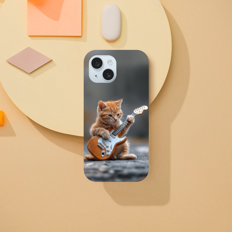 May include: A phone case with a cute orange tabby kitten playing an electric guitar. The case has a dark gray background.