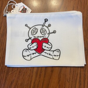 Könnte beinhalten: Weiße Canvas-Reißverschlusstasche mit einer Voodoo-Puppe, die ein rotes Herz hält. Die Puppe hat gestickte Details, Knopfaugen und Nadeln. Ein Etikett mit einem schwarzen Katzenlogo ist angebracht.