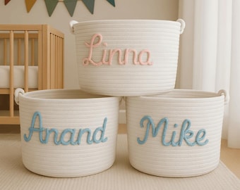 May include: Three white rope storage baskets with handles. The top basket has the name "Linna" in pink script. The bottom two baskets have the names "Anand" and "Mike" in blue script. A crib and a colorful banner are in the background.