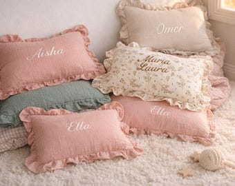 Puede incluir: Una colección de cojines decorativos en varios colores, incluyendo rosa, beige y verde, con bordes con volantes. Algunos cojines tienen nombres bordados como "Aisha", "Omer" y "Ella". Los cojines están colocados sobre una alfombra blanca y esponjosa.
