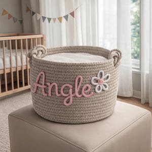 Peut inclure: Un panier en corde beige avec le nom "Angle" en écriture rose et une fleur blanche appliquée. Le panier a deux poignées en corde et est rempli d'une couverture blanche. Le panier est posé sur un pouf beige.