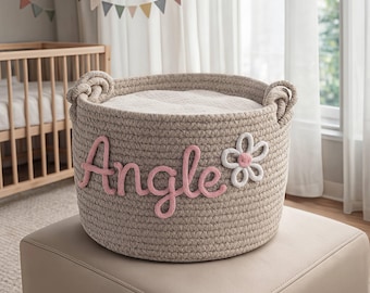 May include: A beige rope basket with the name "Angle" in pink script and a white flower applique. The basket has two rope handles and is filled with a white blanket. The basket is sitting on a beige ottoman.