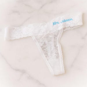 May include: White lace thong underwear with the text "Mrs. Johnson" embroidered in light blue. The delicate lingerie features a scalloped lace trim and a small bow detail. The thong is designed with a comfortable fit and a classic style.