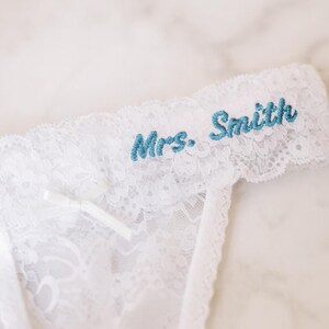May include: White lace garter with the embroidered text "Mrs. Smith" in teal. The garter features a delicate floral lace pattern and a small white bow. A bridal accessory.