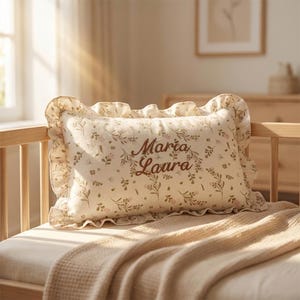 Puede incluir: Una almohada rectangular con un estampado floral y bordes con volantes, apoyada sobre una cama. La almohada presenta los nombres bordados "Maria Laura" en una fuente marrón. La funda de almohada tiene un fondo crema con detalles florales rosas y verdes.