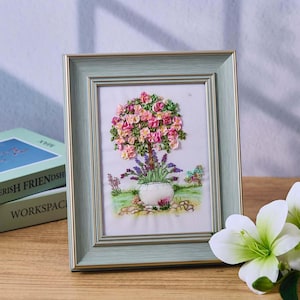 May include: A framed embroidery piece depicting a pink and white flower tree in a white pot with lavender flowers at the base. The embroidery is on a white background and is framed in a light green frame.