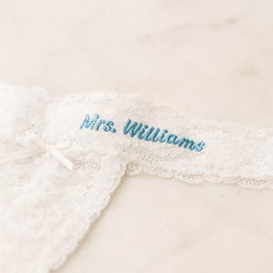 May include: White lace garter with a delicate bow and the embroidered text "Mrs. Williams" in light blue. The garter features a scalloped edge and is designed for a wedding.