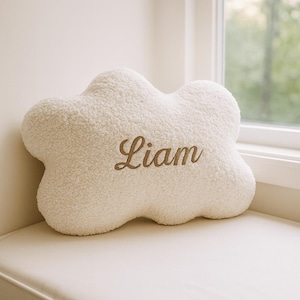 May include: A cloud-shaped cushion in a soft, off-white hue. The cushion has a textured, plush surface and features the name "Liam" embroidered in a brown script font. The cushion is placed on a white surface near a window.