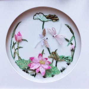 May include: A framed, circular artwork featuring embroidered pink lotus flowers, buds, and green leaves with sequins. Two dragonfly figures are also embroidered. The artwork is set against a white background.