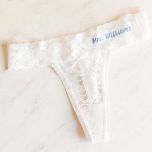 May include: White lace thong underwear with the text "Mrs. Williams" in blue script. The delicate lingerie features a floral lace pattern and scalloped edges. A bridal or lingerie item.