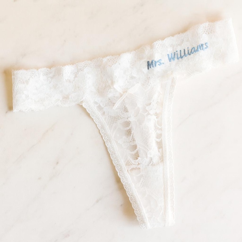 May include: White lace thong underwear with the text "Mrs. Williams" in blue script. The delicate lingerie features a floral lace pattern and scalloped edges. A bridal or lingerie item.