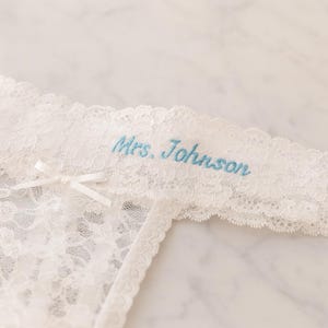 May include: Ivory lace lingerie with a small bow and the embroidered text "Mrs. Johnson" in pale blue. The lingerie has a scalloped edge and detailed floral lace.