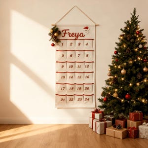 May include: A festive cream advent calendar with red pockets numbered 1 to 24, with the name "Freya" at the top. A Christmas tree with lights and baubles, and wrapped gifts are in the background, creating a holiday scene.