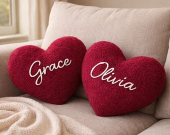 May include: Two heart-shaped burgundy pillows with white embroidered names, "Grace" and "Olivia". The pillows are on a beige couch with a cream-colored blanket. The pillows have a textured, plush appearance.