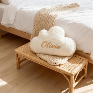 May include: A fluffy, cloud-shaped pillow in cream color, embroidered with the name "Olivia" in gold thread. The pillow rests on a small, woven wicker bench, alongside a matching cream-colored knitted throw. The scene is set in a bedroom.