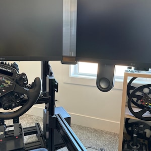 Puede incluir: Una configuración de simulador de carreras negra con dos monitores LG. La configuración incluye un volante, pedales y un asiento. Volantes adicionales y un joystick son visibles en una estantería de madera. La habitación tiene una ventana y una alfombra gris.