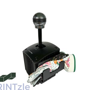 Puede incluir: Palanca de cambios Fanatec negra con pomo plateado, junto con guantes de carreras con un colorido diseño floral y geométrico. Los guantes se sujetan en un soporte negro fijado a la palanca.