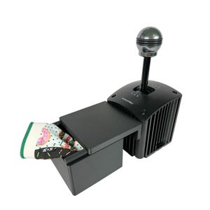 May include: A black and silver manual gear shifter with a black box with a lid that is open and a patterned fabric inside. The gear shifter has a silver knob and a black base with a silver plate that says "FANATEC".