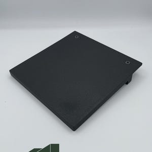 Puede incluir: Plataforma cuadrada negra con bordes redondeados y dos tornillos visibles. La plataforma tiene una superficie texturizada y un pequeño labio en un lado. El logotipo "TPRINTzle" es visible en la esquina inferior izquierda.